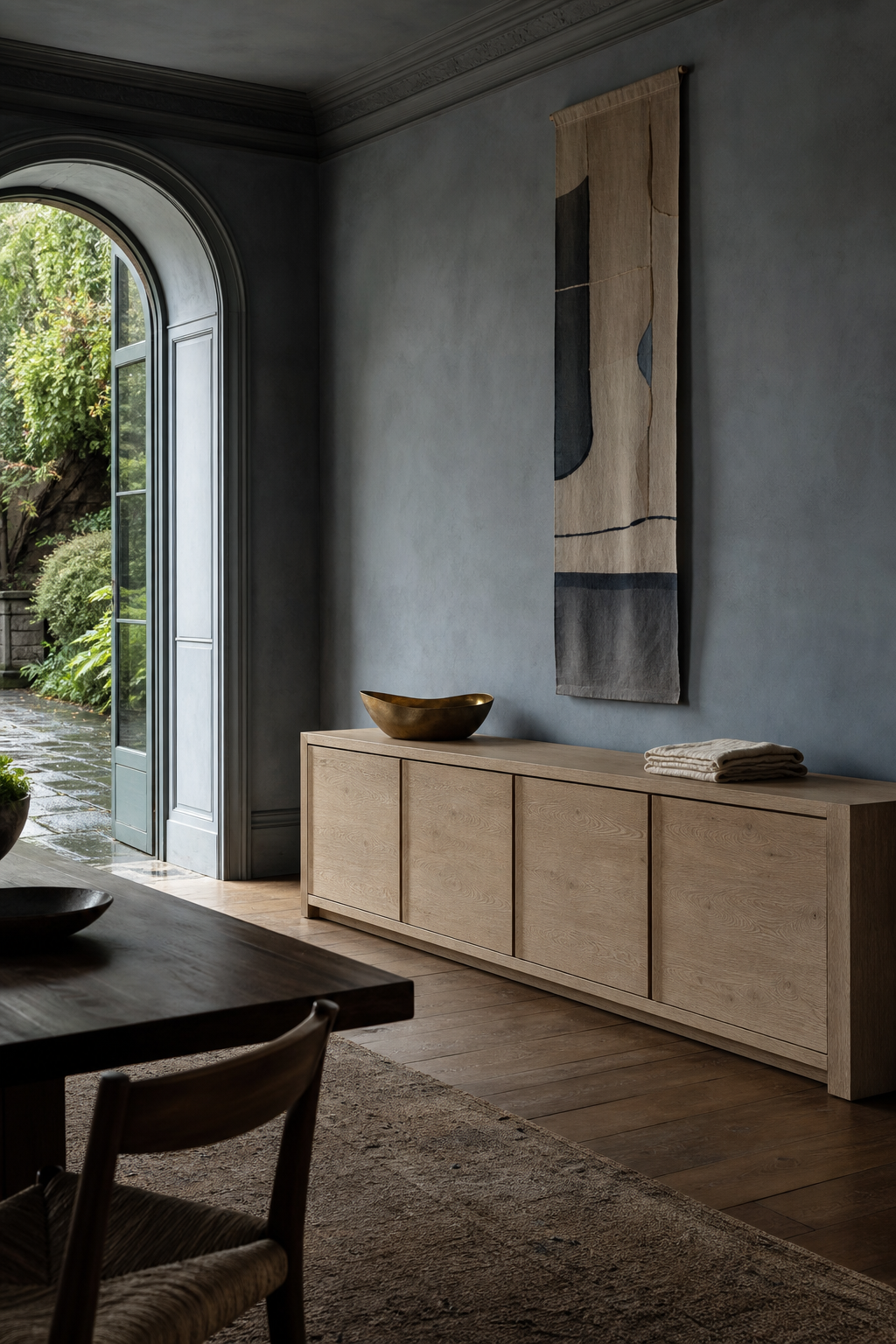 Conceptual AI-modified mineral-blue dining room organized around a long pale sideboard with clear circulation in front and a rain-dark garden beyond